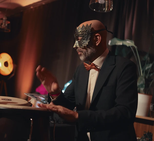 En mann iført dress og maskeradeballmaske spiller trommer. Foto.